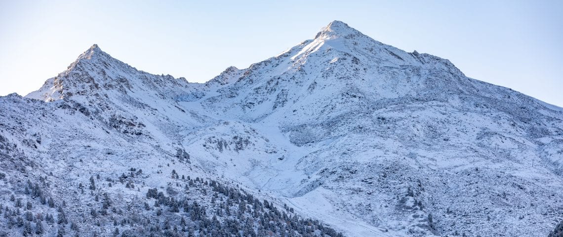 Mountains covered in snow with the sun behind them.